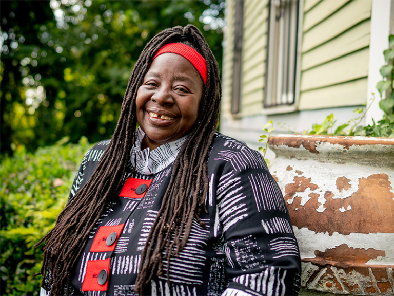 a beautiful black woman with long dreadlocks, a blue and white patterned top, and a red headband smiles at the camera against a backdrop of a building and trees