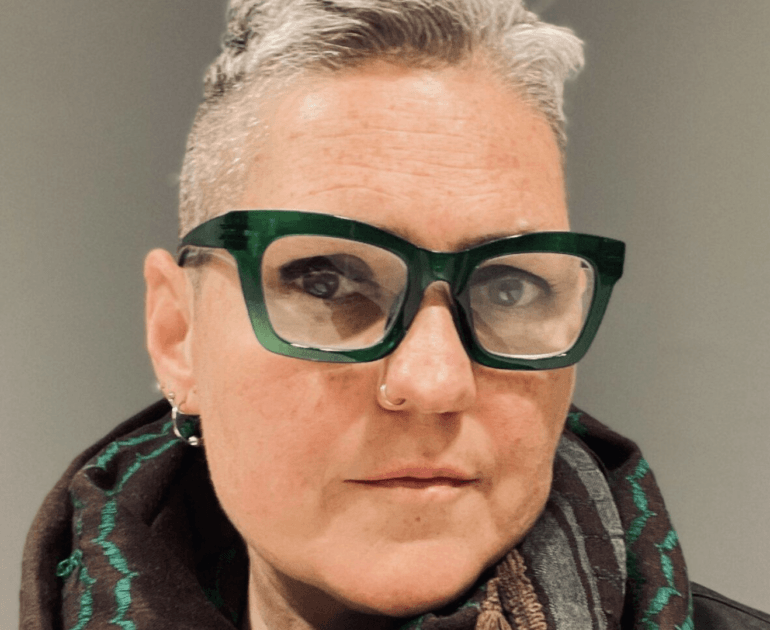 headshot of a middle aged white person with big blocky glasses, a blonde buzz cut, and earrings. She wears a green and gray scarf and stares into the camera.