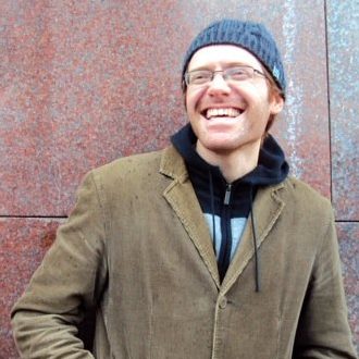 a white person with reddish hair and a gray beanie + greenish khaki jacket smiles big and leans against a rust-red wall.